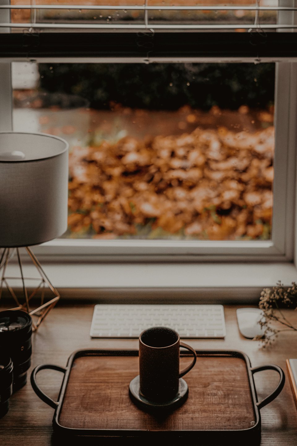 A desk with a keyboard, mug, and lamp near a window.