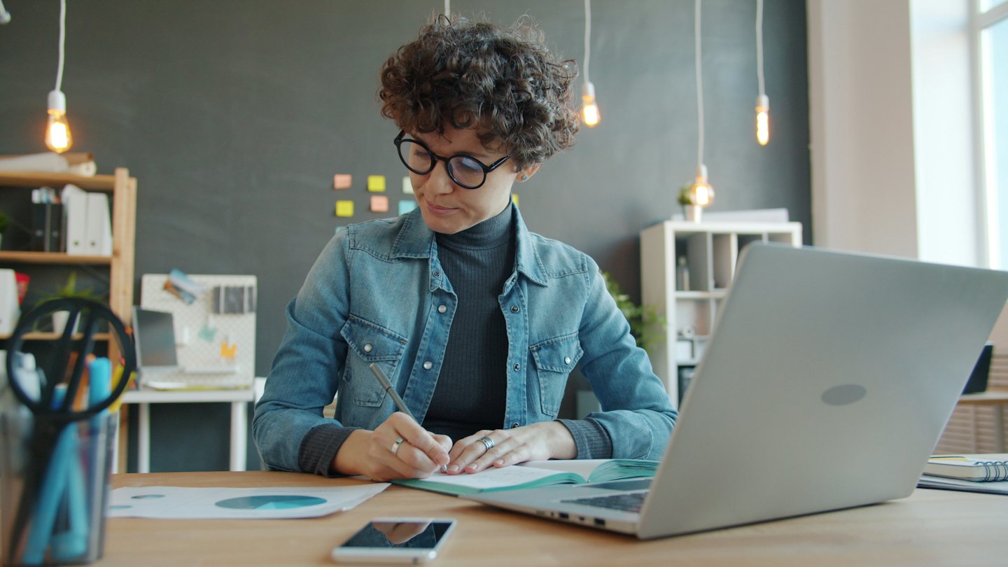 Woman with curly hair writing in notebook at desk with laptop.