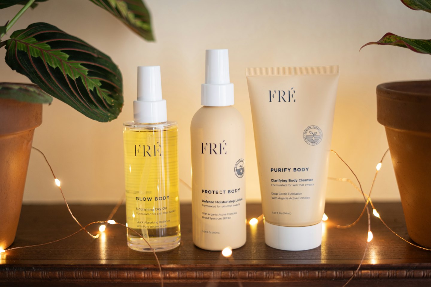 Three FRÉ skincare products on dark wood with plants and fairy lights.